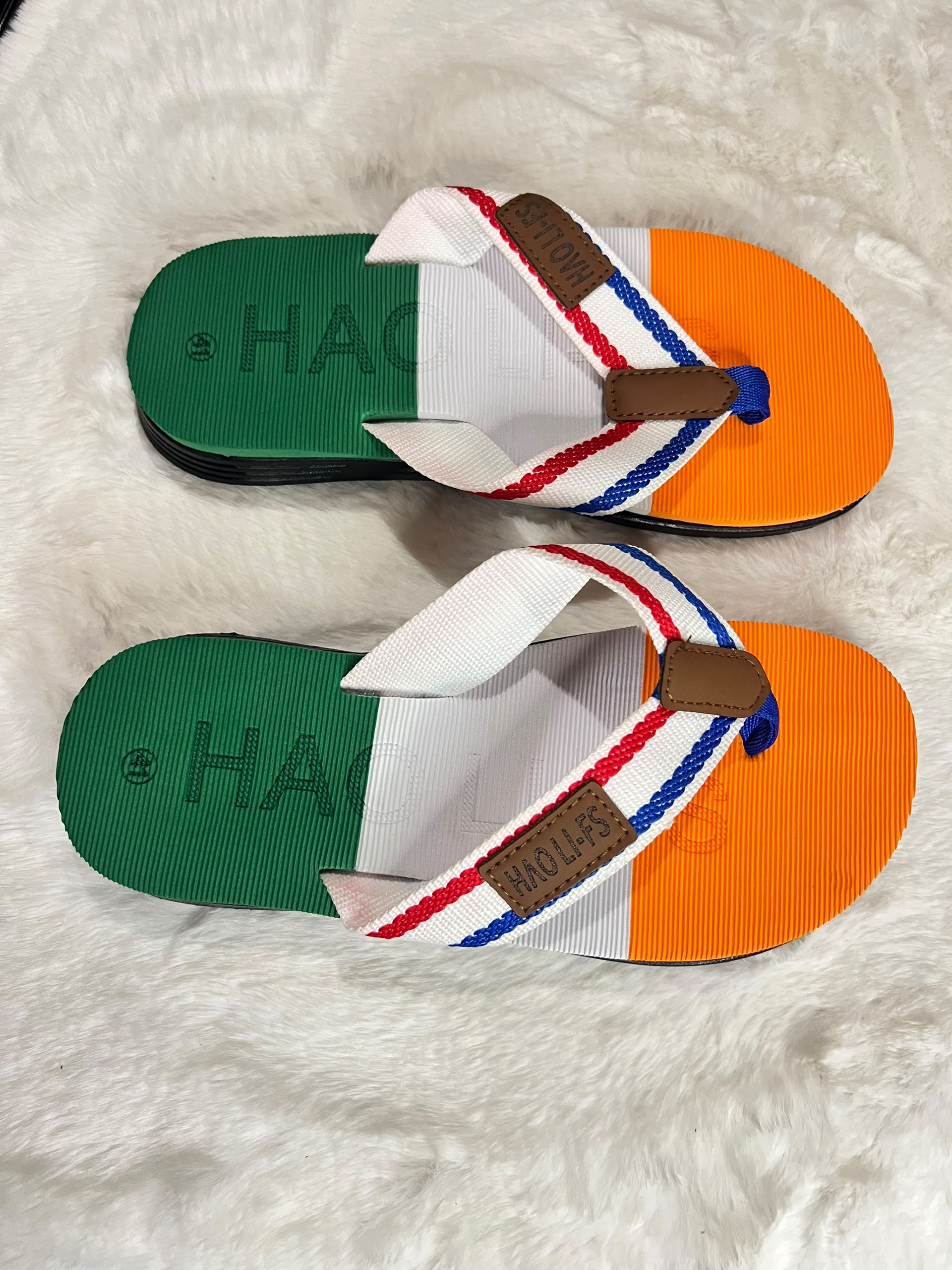 A pair of Hágás flip-flop sandals with a green, white, and orange color-blocked design, featuring a white strap with red, white, and blue stripes and a brown brand patch, displayed on a white faux fur background.