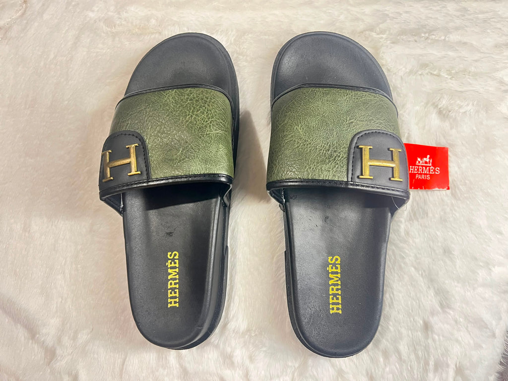 A pair of Hermès slide sandals with olive green textured straps and black soles, featuring a gold 'H' logo on the strap and 'HERMÈS' printed in gold on the footbed. A red Hermès tag is attached to one sandal.