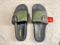 A pair of Hermès slide sandals with olive green textured straps and black soles, featuring a gold 'H' logo on the strap and 'HERMÈS' printed in gold on the footbed. A red Hermès tag is attached to one sandal.
