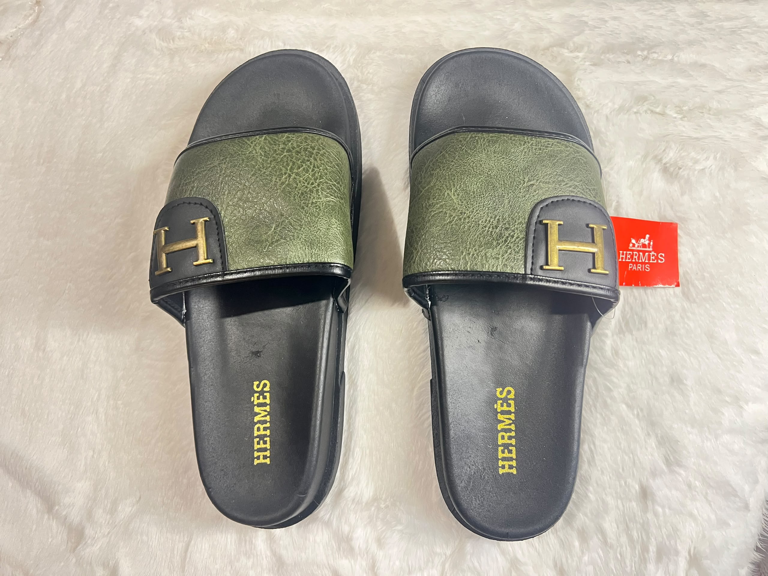 A pair of Hermès slide sandals with olive green textured straps and black soles, featuring a gold 'H' logo on the strap and 'HERMÈS' printed in gold on the footbed. A red Hermès tag is attached to one sandal.
