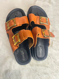 A pair of women's double-strap slide sandals in tan leather with gold-tone buckles and black soles, displayed on a white faux fur surface. The brand name 'coolgirl' is visible on the footbed.