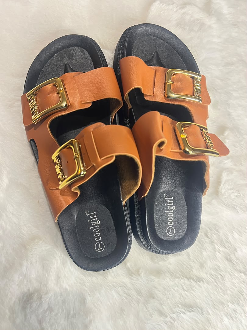 A pair of women's double-strap slide sandals in tan leather with gold-tone buckles and black soles, displayed on a white faux fur surface. The brand name 'coolgirl' is visible on the footbed.