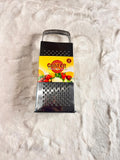 A stainless steel box grater with a black handle and a yellow brand label reading 'Crater' lies on a white faux fur surface.