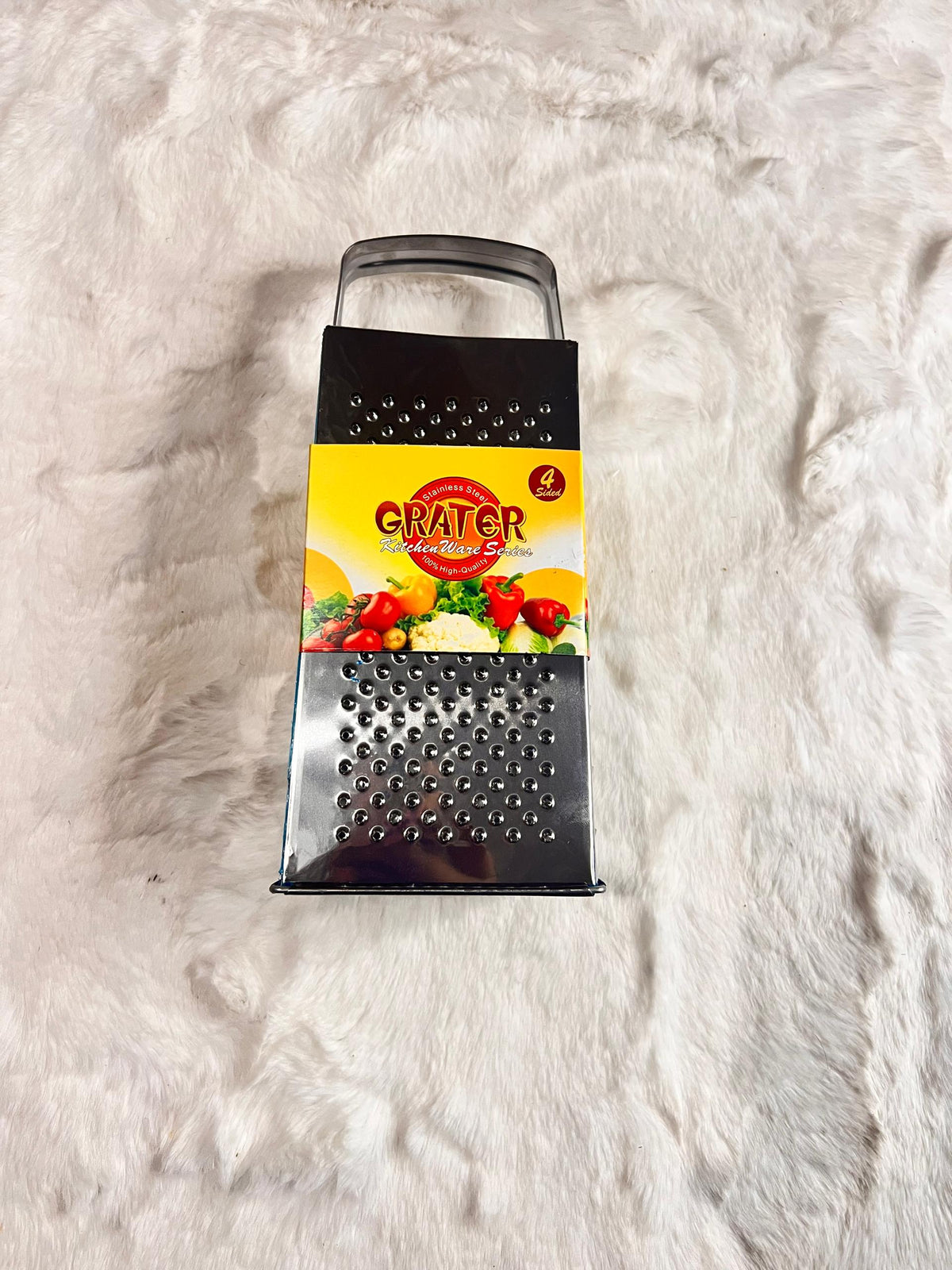 A stainless steel box grater with a black handle and a yellow brand label reading 'Crater' lies on a white faux fur surface.