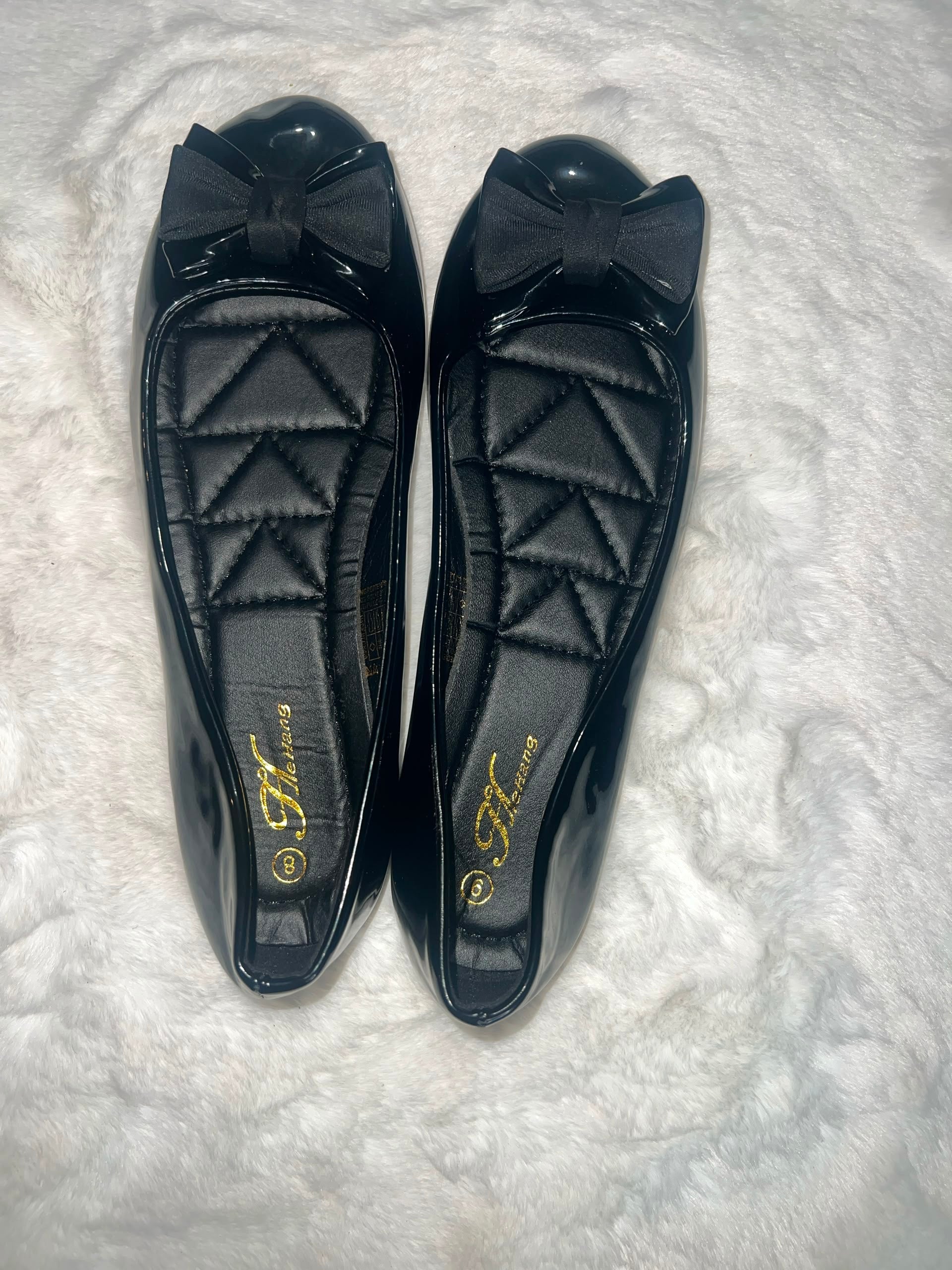 A pair of black patent leather ballet flats with quilted insoles and a bow on the toe, displayed on a white textured faux fur surface. The insole features a gold 'Fashion' logo.