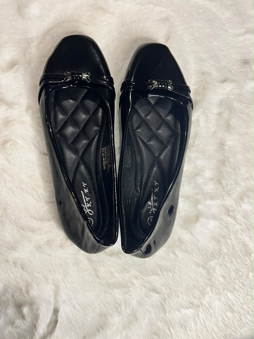 A pair of black quilted leather ballet flats with metallic silver bow details on the toes, displayed on a white faux fur surface.