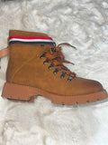 A brown suede lace-up ankle boot with a thick lug sole, resting on a white textured faux fur surface. The collar features a red, white, and navy blue striped band.