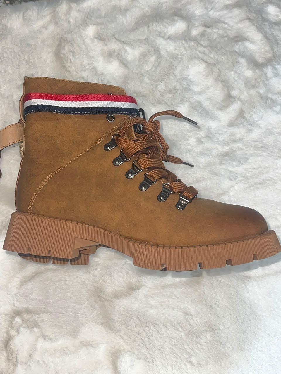 A brown suede lace-up ankle boot with a thick lug sole, resting on a white textured faux fur surface. The collar features a red, white, and navy blue striped band.