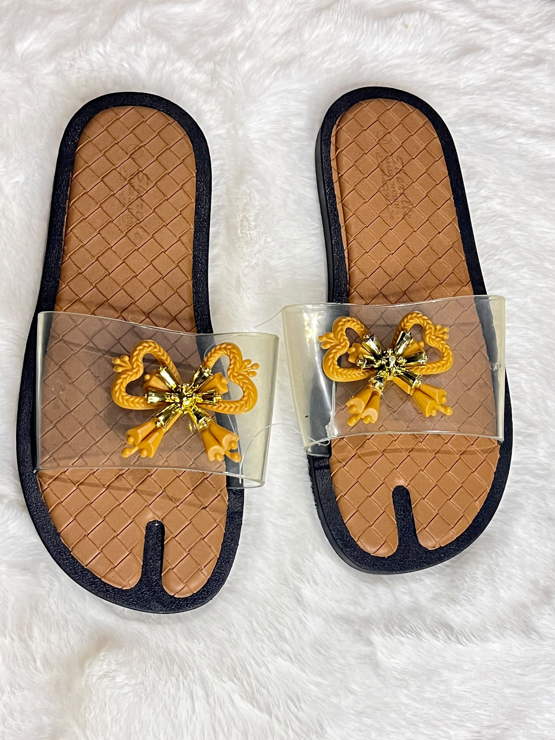A pair of tabi-style slide sandals with tan woven insoles, black trim, clear plastic straps, and a yellow decorative bow embellishment on the front, displayed on a white faux fur surface.
