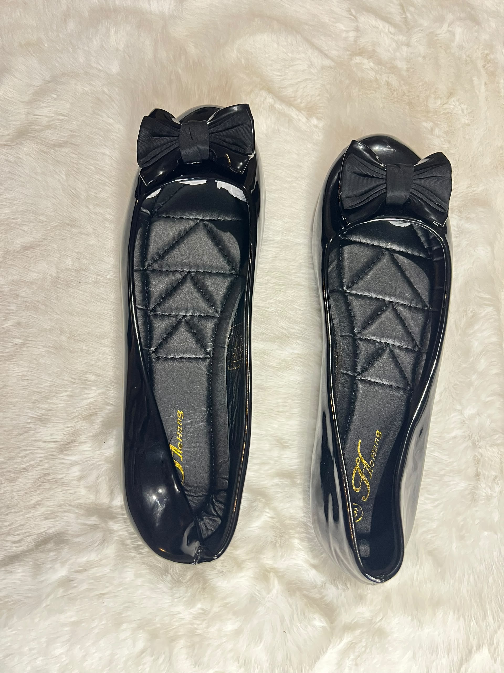 A pair of black patent leather ballet flats with quilted insoles and a bow detail on the toe, displayed on a white faux fur surface.