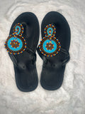 A pair of black thong sandals with intricate hand-beaded straps featuring turquoise blue, yellow, red, and black patterns, laid flat on a white faux fur surface.