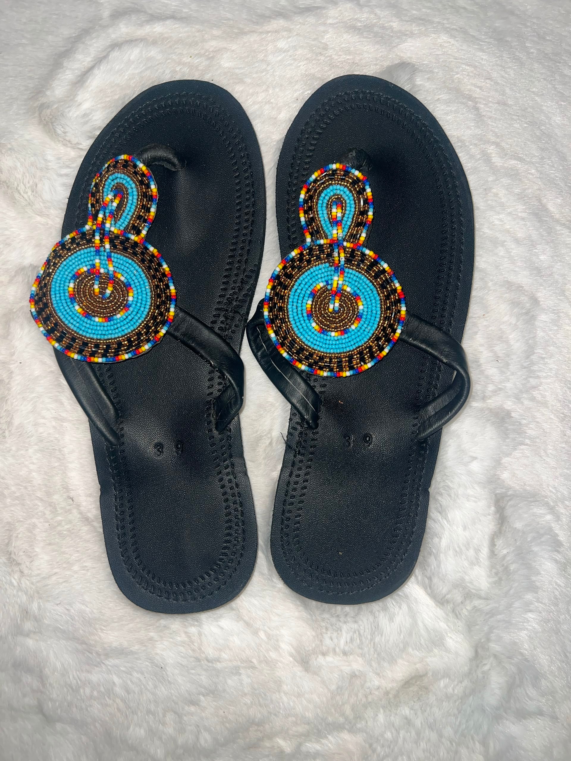 A pair of black thong sandals with intricate hand-beaded straps featuring turquoise blue, yellow, red, and black patterns, laid flat on a white faux fur surface.