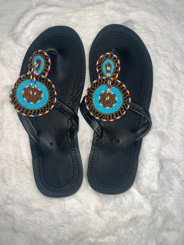 A pair of black thong sandals with intricate hand-beaded straps featuring turquoise blue, yellow, red, and black patterns, laid flat on a white faux fur surface.