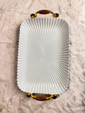 A white rectangular serving tray with a fluted, sunburst pattern and ornate gold handles with brown accents