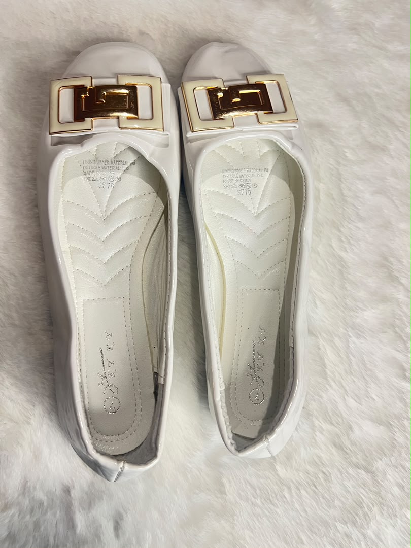 A pair of white leather ballet flats with a prominent gold-tone rectangular buckle detail on the toe, displayed on a white faux fur surface.
