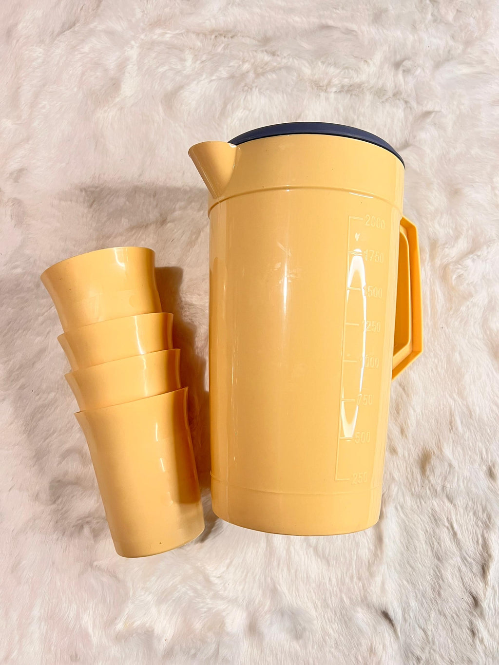 A yellow plastic thermal water bottle with a dark blue lid, shown next to a stack of four matching yellow cups on a white faux fur surface.
