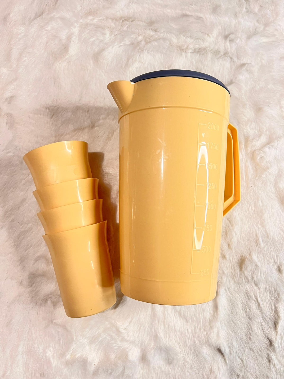 A yellow plastic thermal water bottle with a dark blue lid, shown next to a stack of four matching yellow cups on a white faux fur surface.