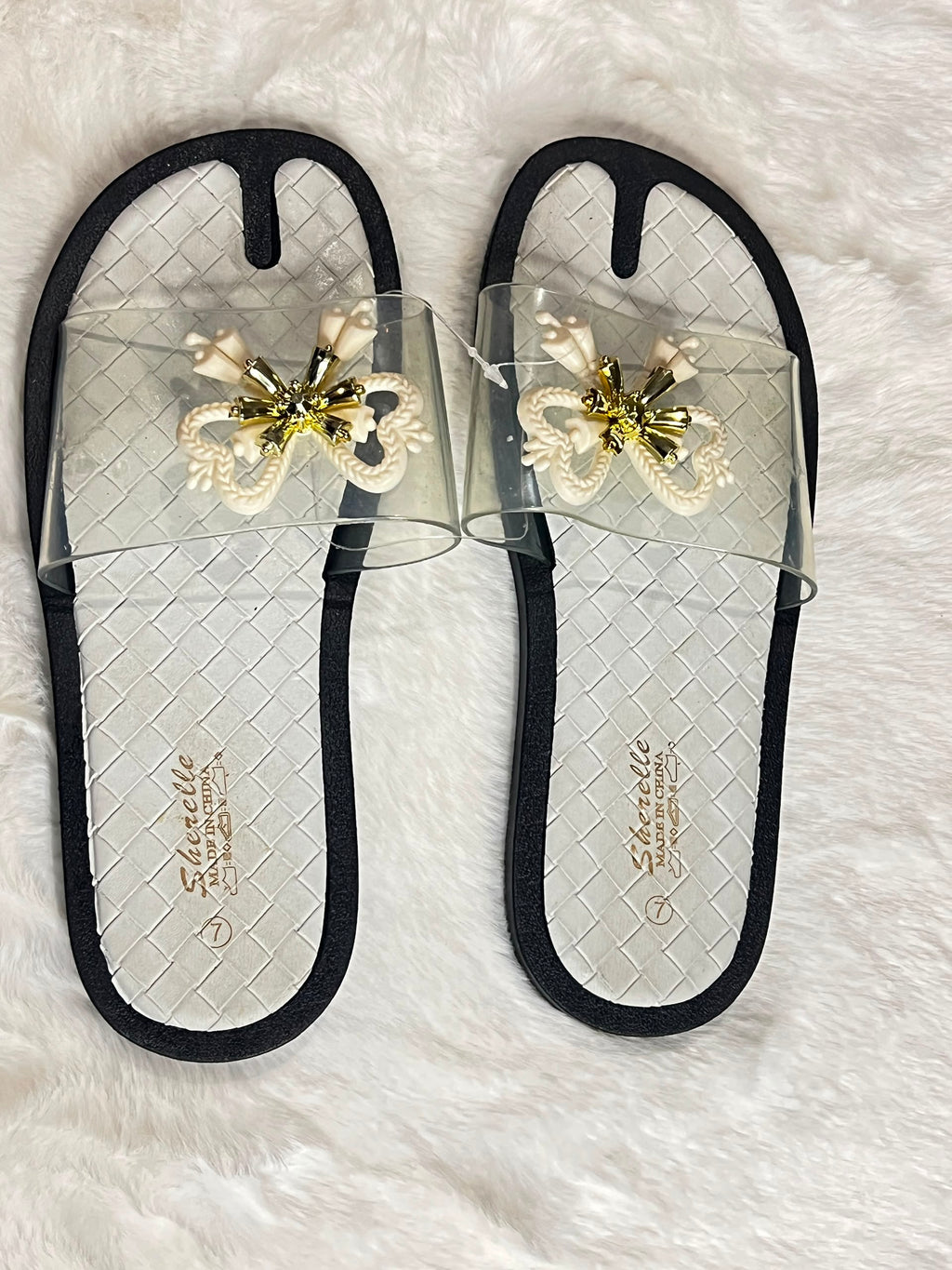 A pair of women's slide sandals with clear mesh straps, black trim, and a decorative white and gold bow embellishment on the top. The insoles feature a black and white woven pattern with the brand name 'Schindello' printed in gold.