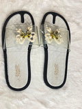 A pair of women's slide sandals with clear mesh straps, black trim, and a decorative white and gold bow embellishment on the top. The insoles feature a black and white woven pattern with the brand name 'Schindello' printed in gold.