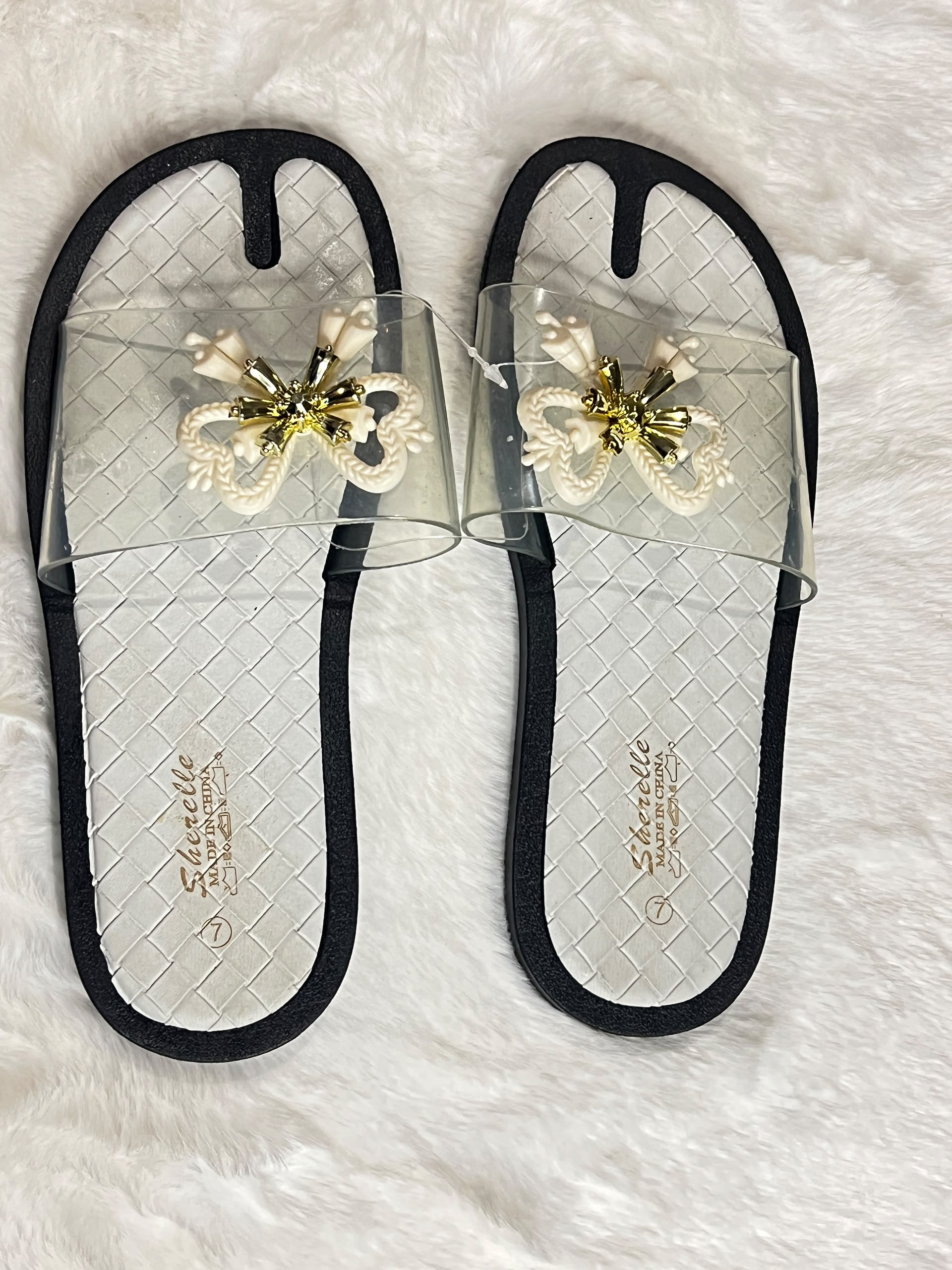 A pair of women's slide sandals with clear mesh straps, black trim, and a decorative white and gold bow embellishment on the top. The insoles feature a black and white woven pattern with the brand name 'Schindello' printed in gold.