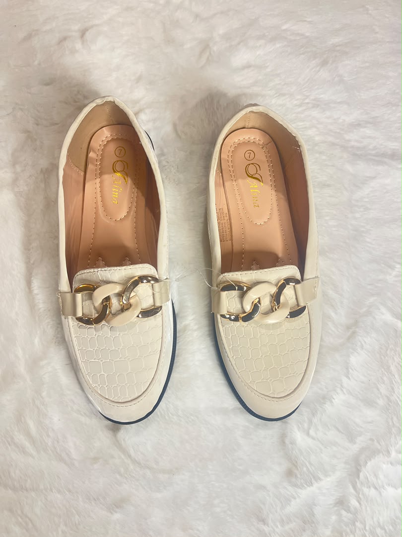 A pair of women's cream-colored loafers with a textured toe cap and a chunky gold-tone chain detail across the vamp, laid flat on a white faux fur surface.