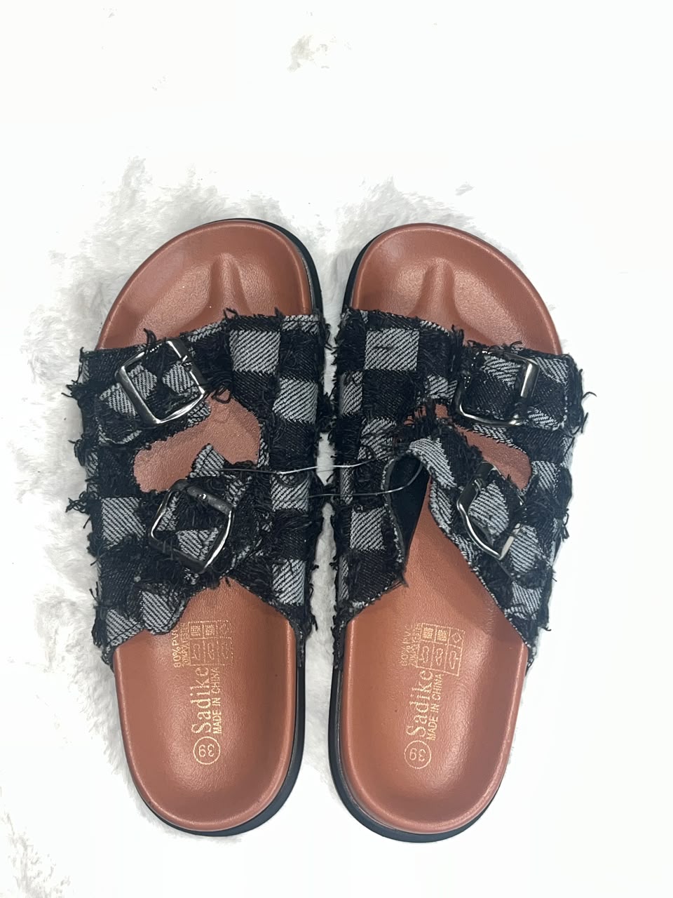 A pair of women's slide sandals with distressed black and gray plaid straps and silver buckle details, set against a white textured background. The brand 'Sodile' is visible on the footbed.