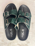 A pair of dark green double-strap slide sandals with silver buckles and black soles, laid flat on a white faux fur surface.