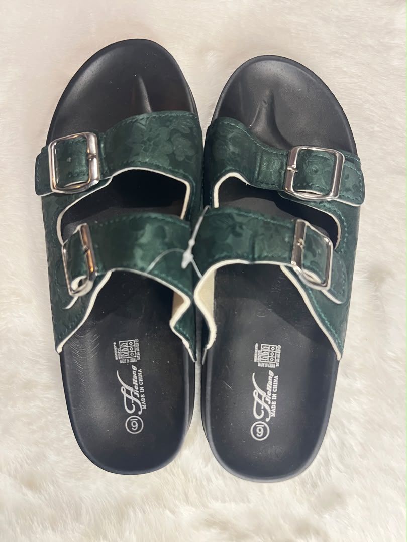 A pair of dark green double-strap slide sandals with silver buckles and black soles, laid flat on a white faux fur surface.