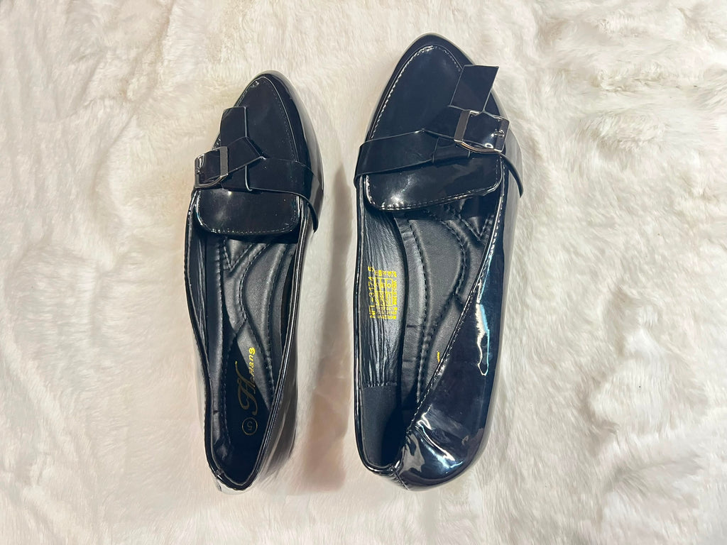 A pair of black patent leather loafers with a bow and buckle detail on the vamp, displayed on a white faux fur surface.