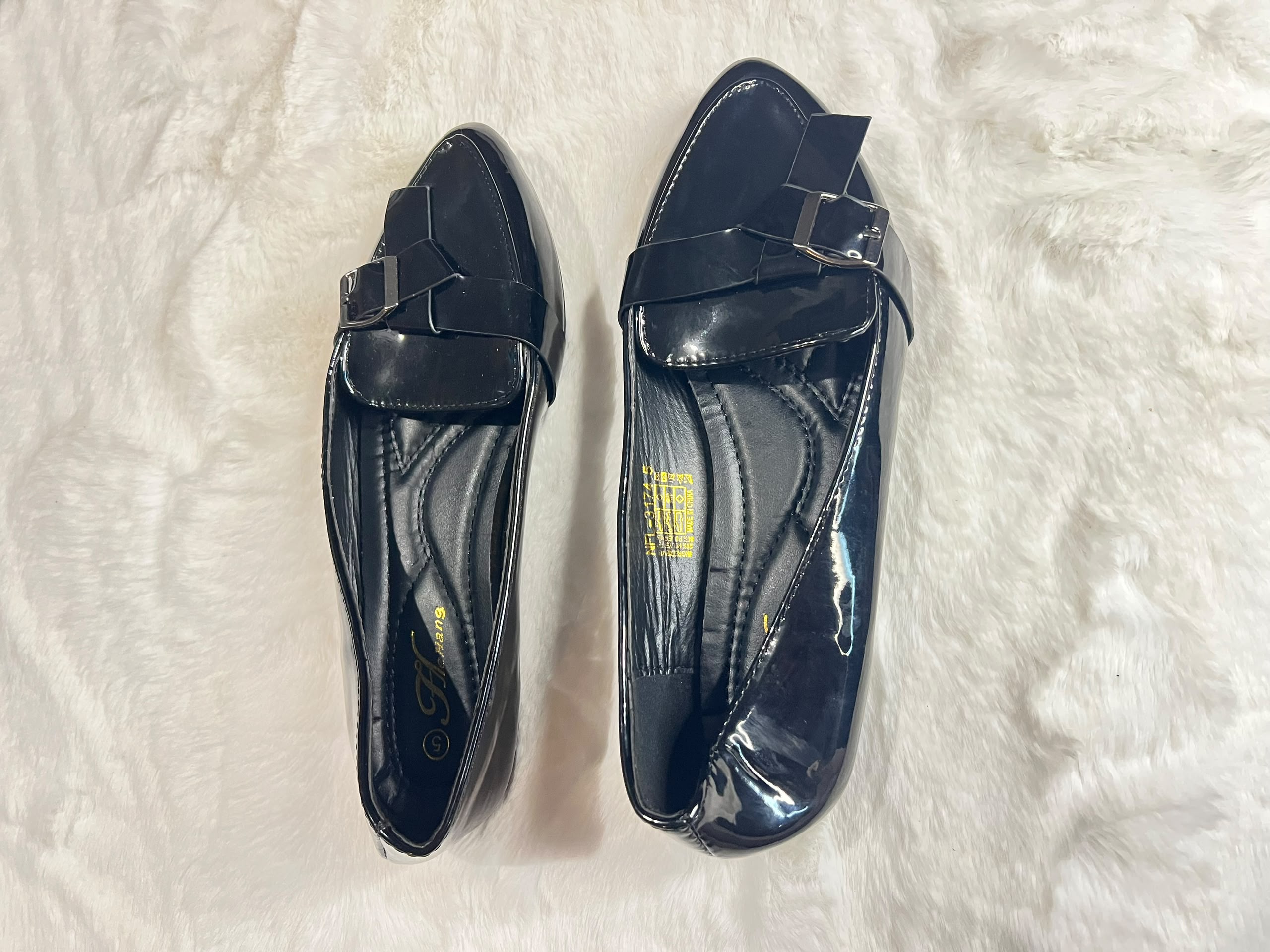 A pair of black patent leather loafers with a bow and buckle detail on the vamp, displayed on a white faux fur surface.