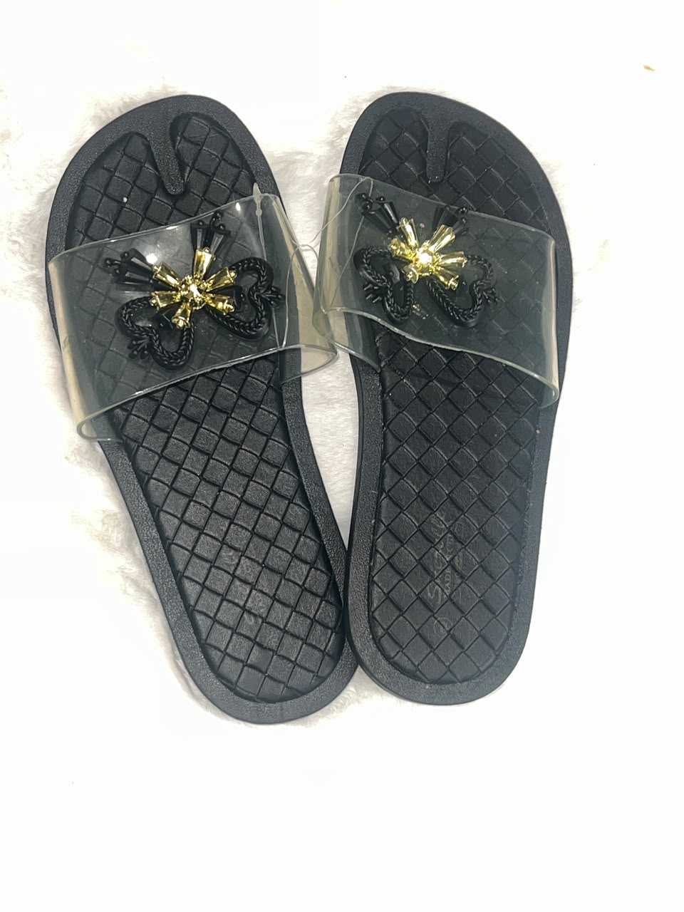 A pair of black woven-texture slide sandals with clear plastic straps decorated with black and gold floral embellishments, shown on a white background.