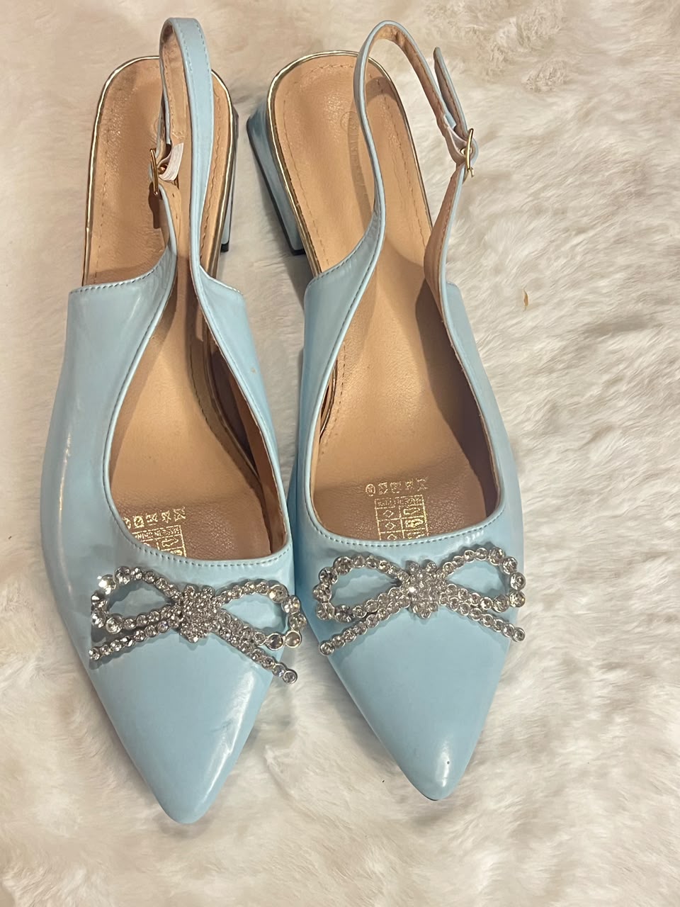 A pair of light blue patent leather slingback pointed-toe flats with silver rhinestone bow embellishments on the toes, displayed on a white faux fur surface.
