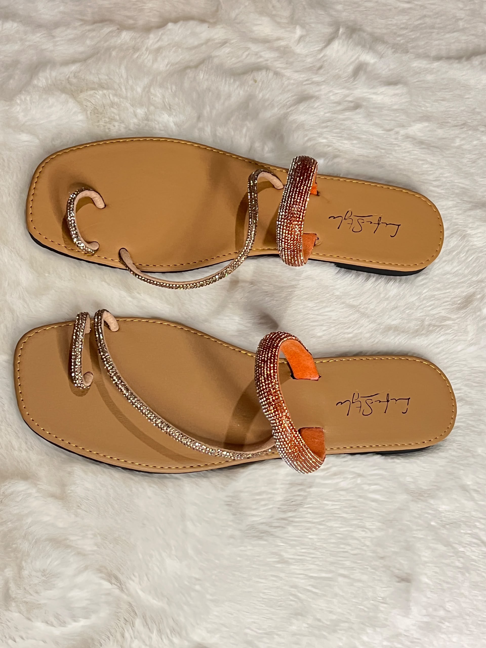 A pair of tan flat thong sandals with rhinestone-embellished toe loops and straps, featuring orange accents on the heel straps, displayed on a white faux fur background.