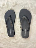 A pair of black flip-flops with glittery metallic straps, displayed flat on a white faux fur surface. The brand name 'BRLA' is printed in white on the sole of each sandal.
