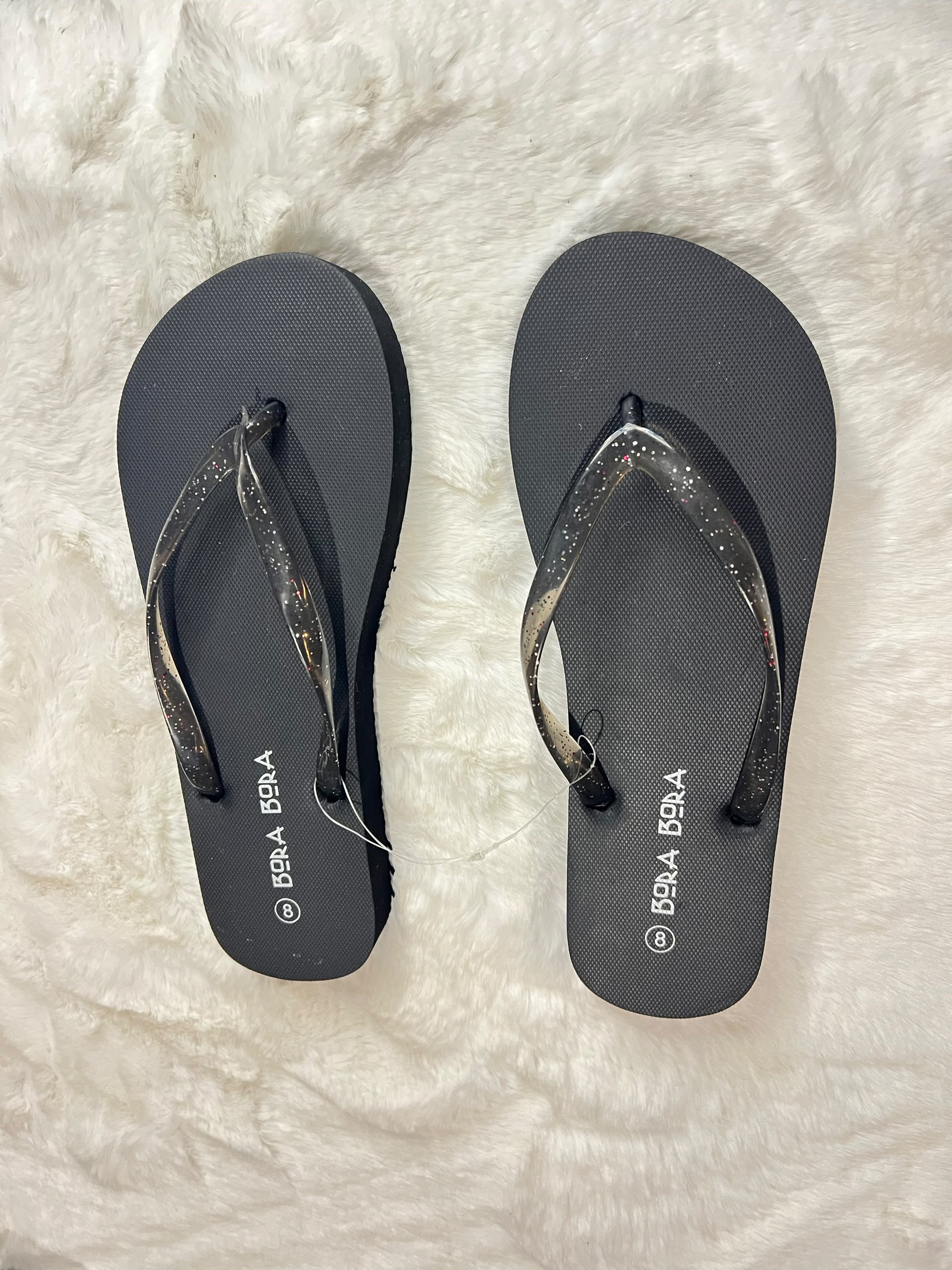 A pair of black flip-flops with glittery metallic straps, displayed flat on a white faux fur surface. The brand name 'BRLA' is printed in white on the sole of each sandal.