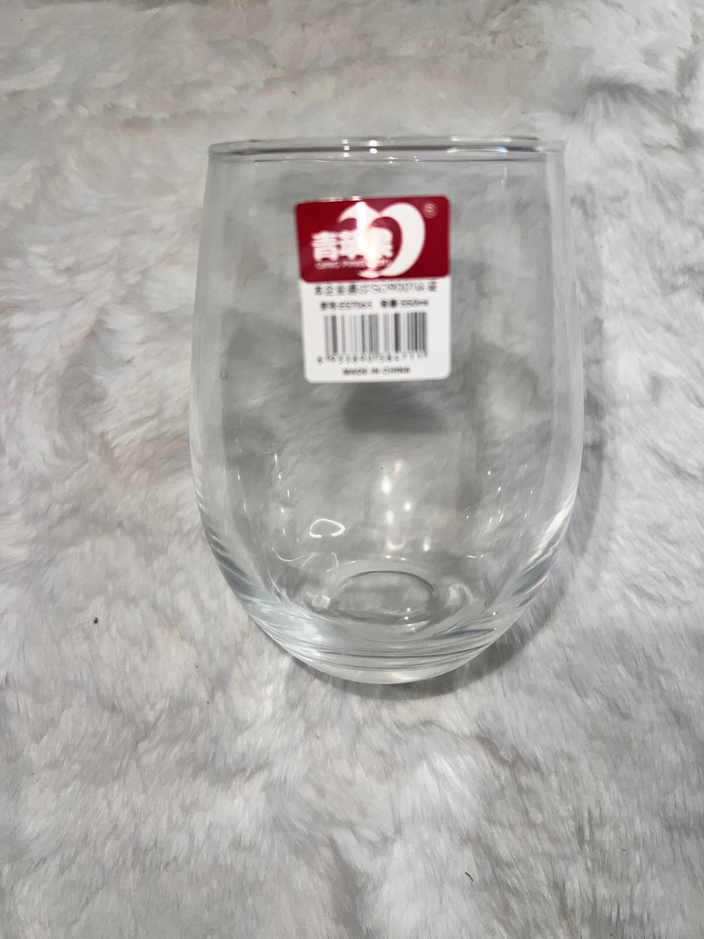 A clear stemless wine glass with a red and white product label sticker on the front, placed on a white faux fur surface.