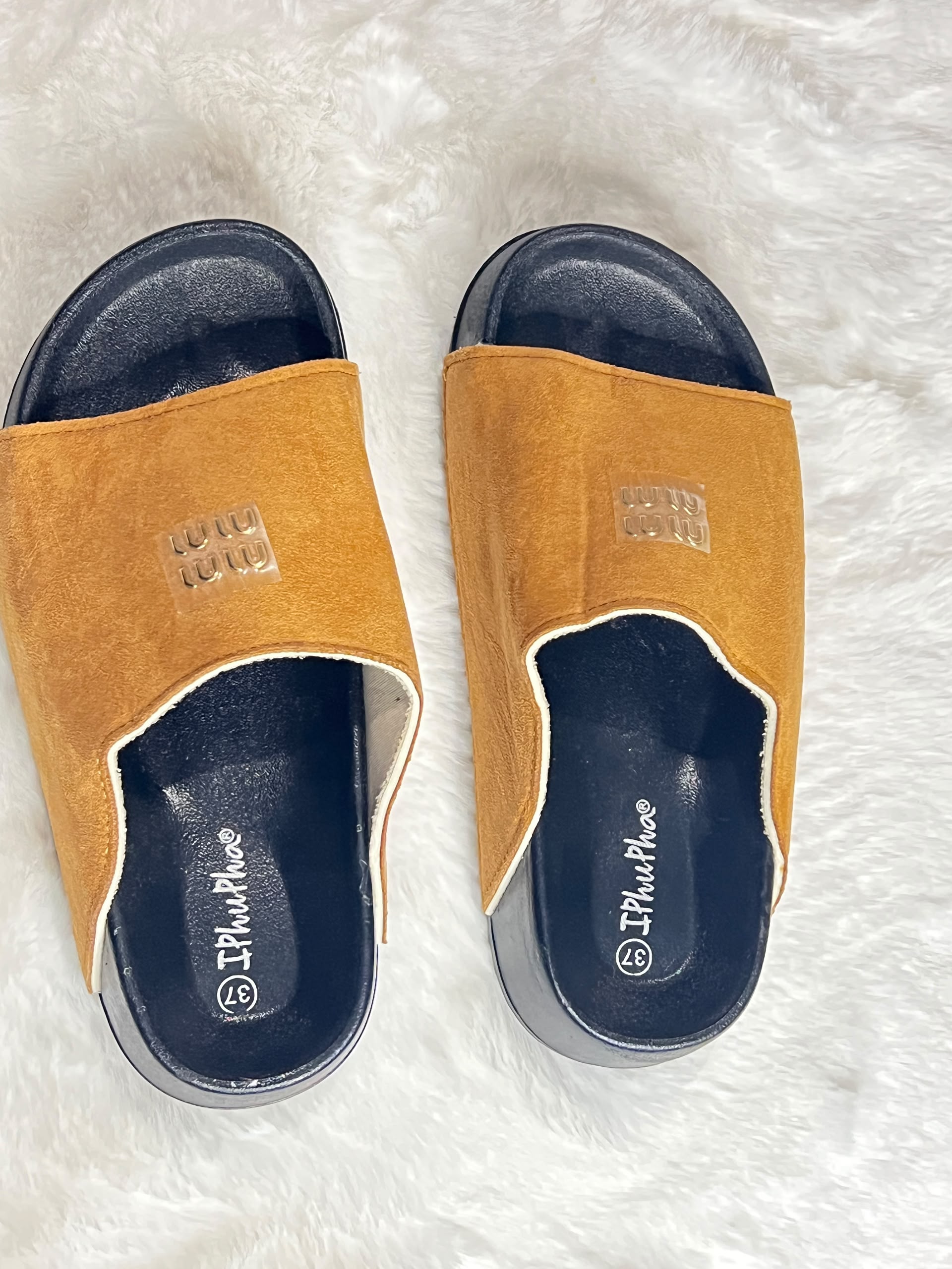 A pair of slide sandals with mustard yellow suede-like uppers and dark navy blue soles, laid flat on a white faux fur surface. The brand name 'IPIfitPhee' is visible on the footbed.