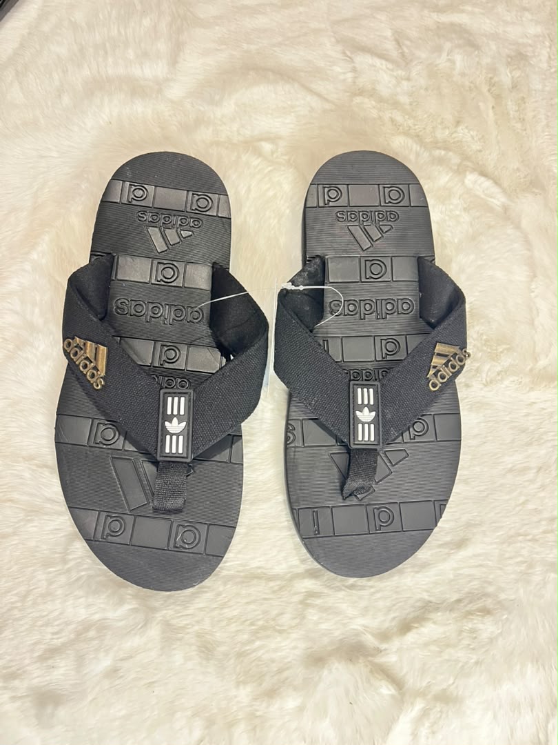 A pair of Adidas flip-flops in dark gray and black, featuring embossed geometric patterns and the Adidas logo on the straps, laid flat on a white faux fur surface.