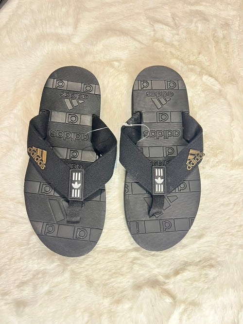 A pair of Adidas flip-flops in dark gray and black, featuring embossed geometric patterns and the Adidas logo on the straps, laid flat on a white faux fur surface.