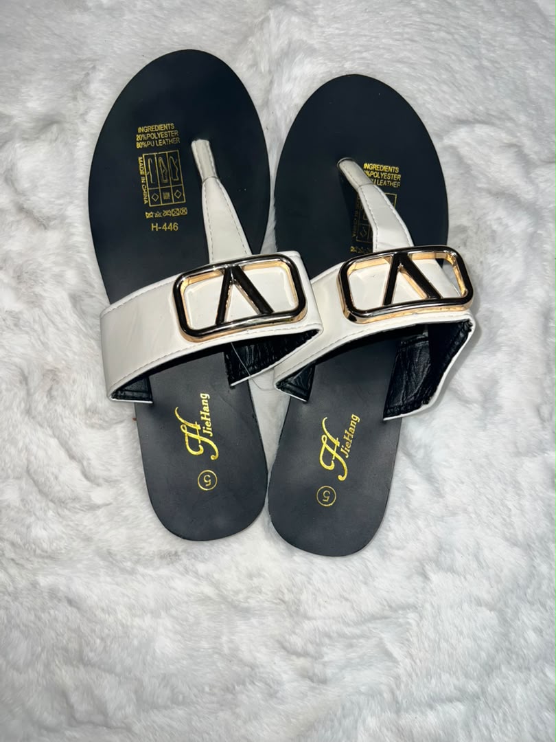 A pair of women's thong sandals with wide white straps featuring large gold and black triangular buckle details, set on a black footbed with gold branding.