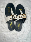 A pair of women's thong sandals with wide white straps featuring large gold and black triangular buckle details, set on a black footbed with gold branding.