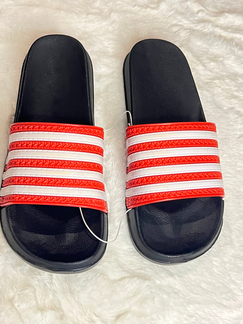 A pair of black slide sandals with red and white striped straps, laid flat on a white faux fur surface.