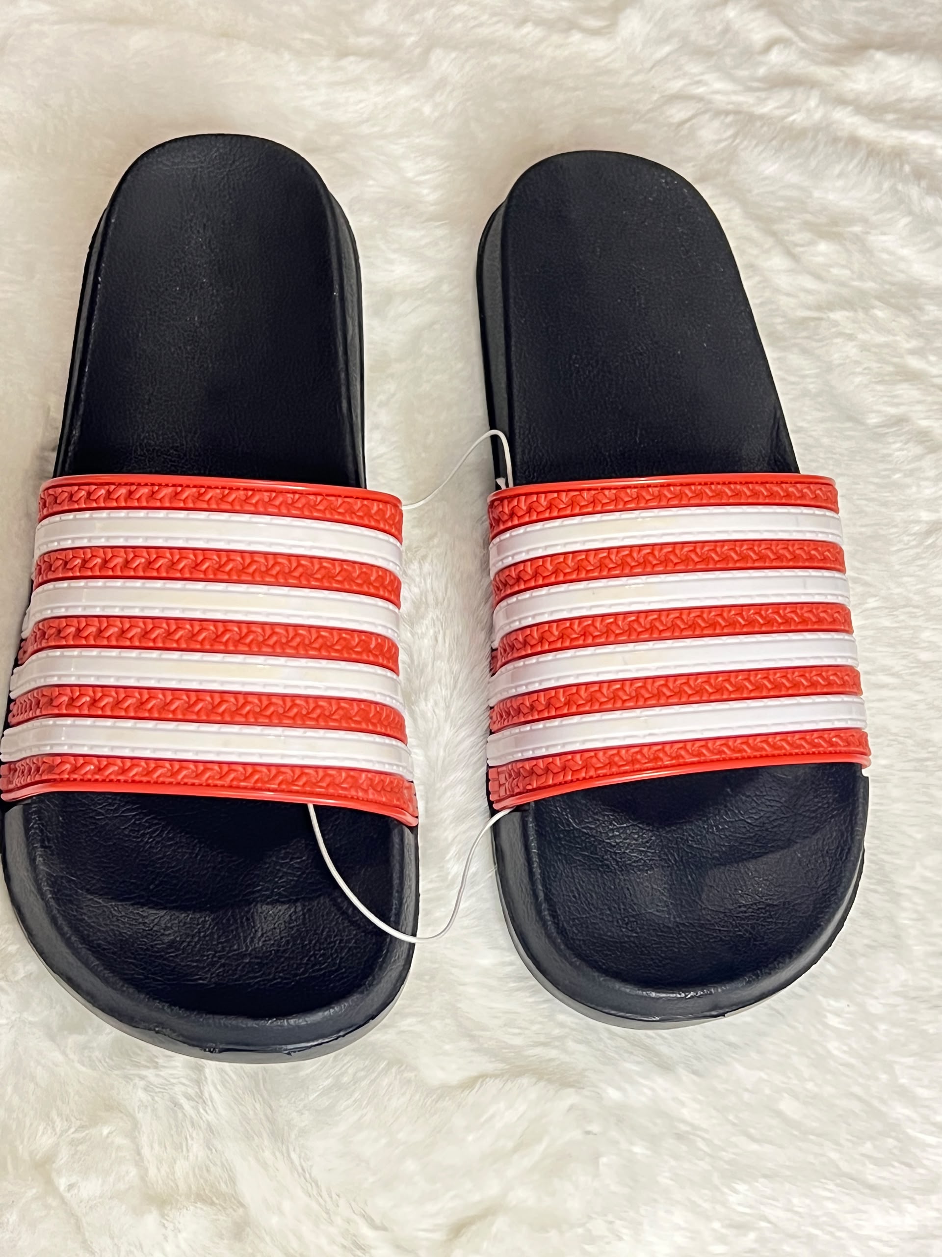 A pair of black slide sandals with red and white striped straps, laid flat on a white faux fur surface.