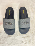 A pair of Zara slide sandals with a textured gray strap featuring a metallic silver-tone ZARA logo, set against a white faux fur background.