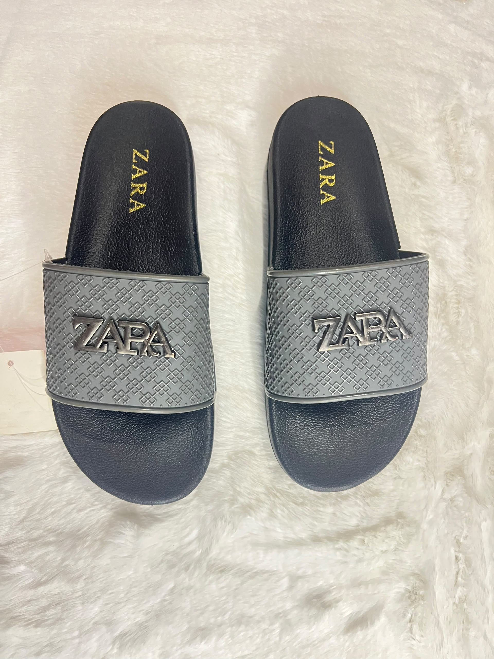 A pair of Zara slide sandals with a textured gray strap featuring a metallic silver-tone ZARA logo, set against a white faux fur background.