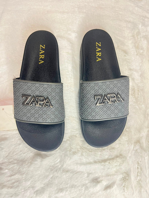 A pair of Zara slide sandals with a textured gray strap featuring a metallic silver-tone ZARA logo, set against a white faux fur background.