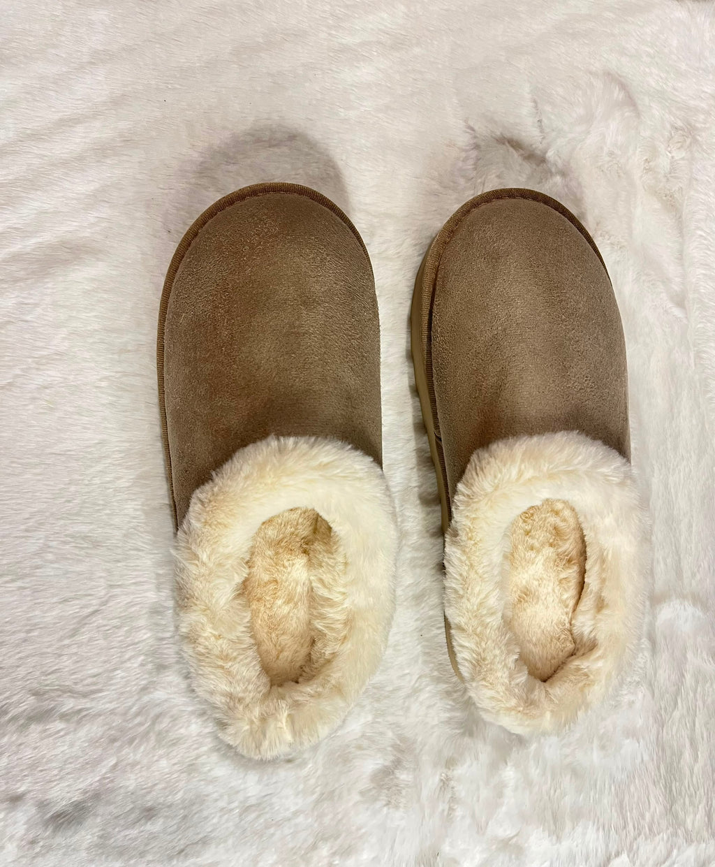 A pair of taupe-brown suede slip-on slippers with thick cream faux-fur trim, laid flat on a white faux-fur rug.