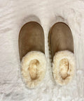 A pair of taupe-brown suede slip-on slippers with thick cream faux-fur trim, laid flat on a white faux-fur rug.