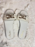 A pair of white slide sandals with metallic gold hardware, displayed on a white faux fur surface. The brand name 'Bruchess' is printed in gold on the footbed.