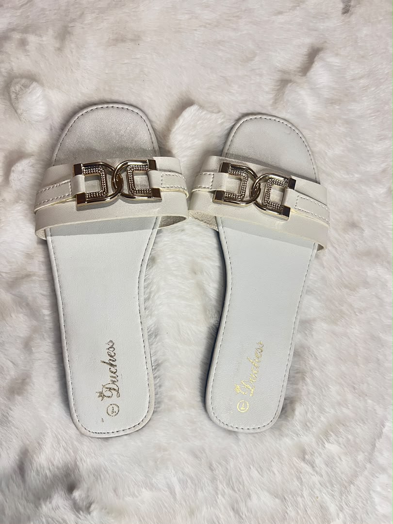 A pair of white slide sandals with metallic gold hardware, displayed on a white faux fur surface. The brand name 'Bruchess' is printed in gold on the footbed.